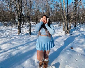 Woman in the woods wearing earmuffs, a pleated blue skirt and brown boots.