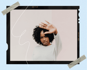 A woman with her hand up, wearing a gray sweatshirt against a beige background.